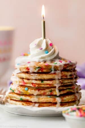 stack of birthday cake funfetti pancakes with icing dripping down the sides, whipped cream on top, and a lit gold candle on top.