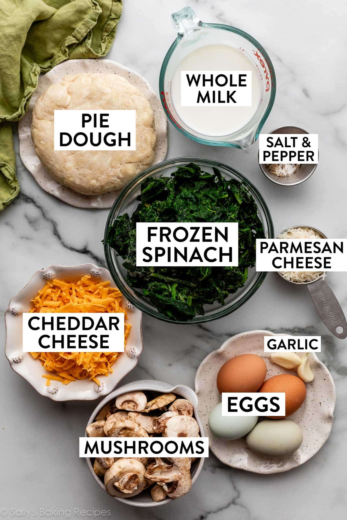 ingredients measured in bowls on marble counter including milk, cheese, eggs, garlic cloves, mushrooms, and spinach.