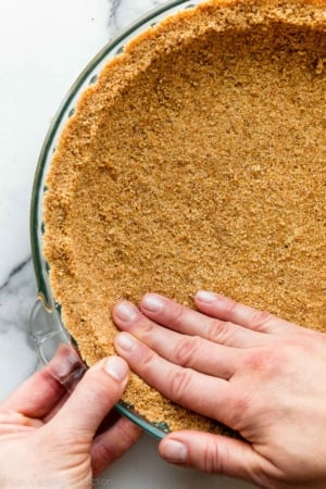 two hands pressing graham cracker crust into glass pie dish.