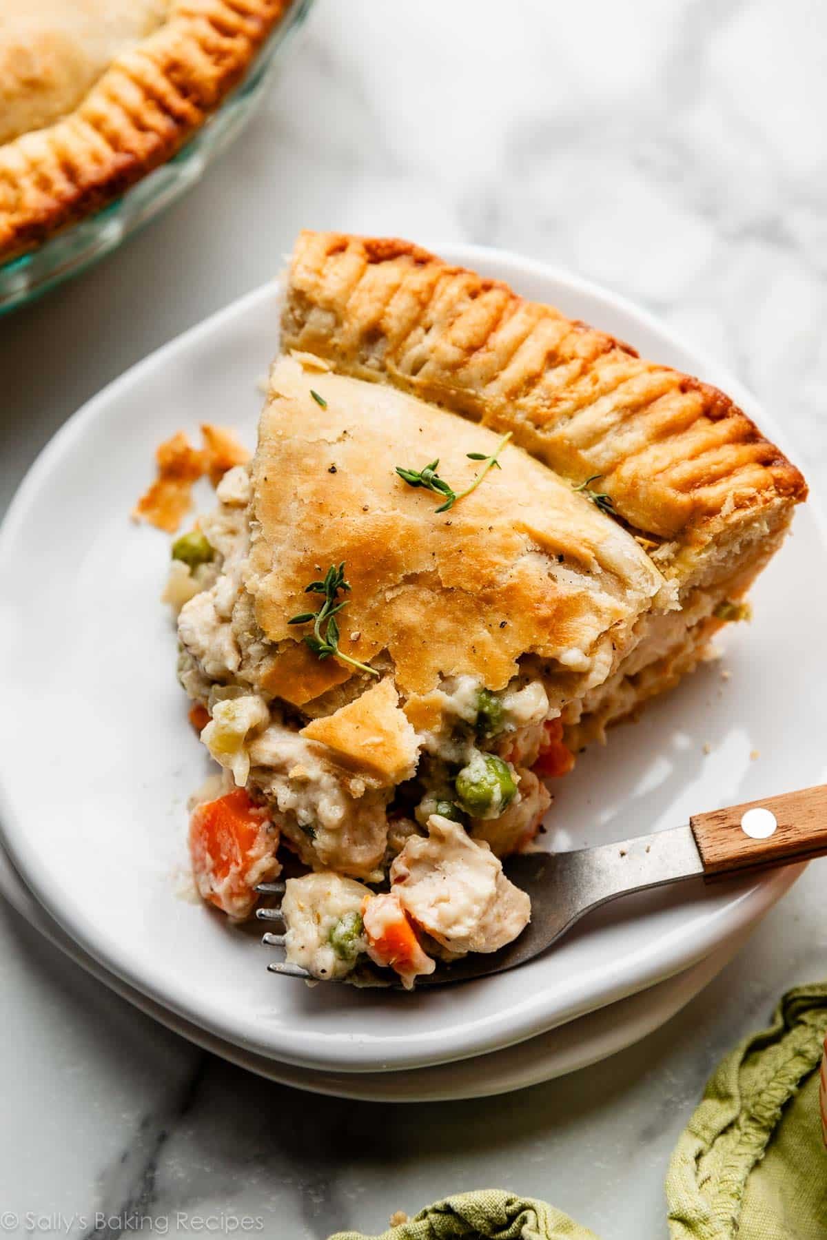 slice of double crust chicken pot pie with golden flaky pie crust on white plate with fork that has a wooden handle.